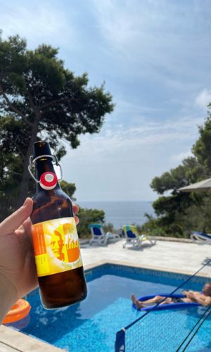 Eine Hand die das 12er Brew Hoppy Summer hält. Im Hintergrund ist ein Swimmingpool in dem ein Mann auf einer Luftmatratze sein Bier genießt. Dahinter ein Blick auf das Meer zwischen Bäumen.