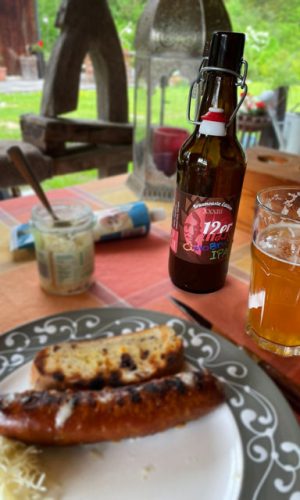 Eine Bierflasche und ein voll gefülltes Glas stehen mit einem Teller auf einem Gartentisch. Auf dem Teller liegt eine Wurst, Brot und Kren.